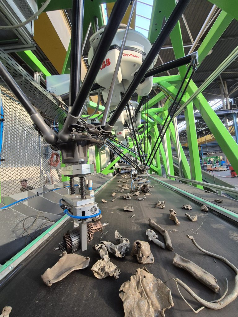Crushed copper motor cores mixed with shredded metal on a conveyor belt under a delta robot
